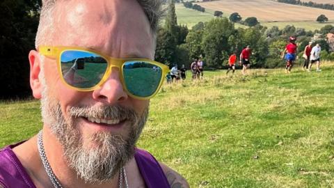 Close-up shot of Sgt Templeman with a beard and moustache wearing yellow-rimmed sunglasses and a purple running top. In the background people can be seen running up a hill with trees in the distance 