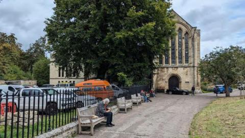 A historic stone church or chapel stands beside a tree-lined pathway, partially shaded by a large mature tree.

Several benches line the path, with a few people seated, while parked vehicles are visible behind a black iron fence.

The scene has a calm, quiet atmosphere under an overcast sky, combining historic architecture with a green outdoor setting.