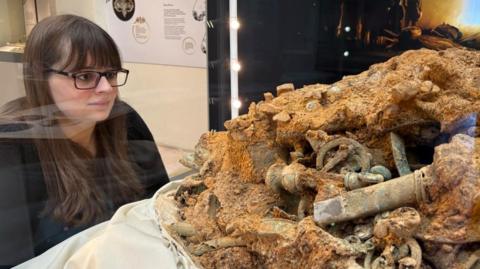 A woman, Emily North, looks into a glass case at a brown-golden mass of metal objects, fused together.