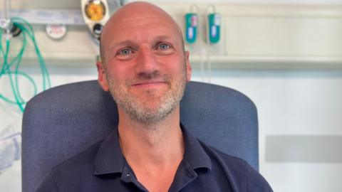 A man with a shaved head and a short grey beard, wearing a dark blue polo shirt, sits in a blue chair, smiling at the camera. Behind him is medical equipment.