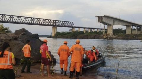 Rescuers stand on the riverbank while others arrive in a boat. In the background is the ruined bridge and another intact bridge further away