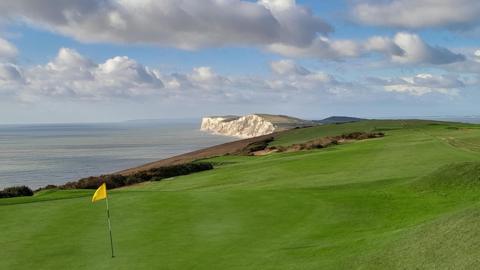 A lush green golf course stretches almost to a cliff edge. Beyond that is the sea and the white cliffs of the coastline can be seen in the distance. In the foreground there is a yellow flag marking a hole on the golf course. 