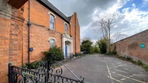 Image shows the side of a red brick house with a blue door and a large tarmac area in front of it. 