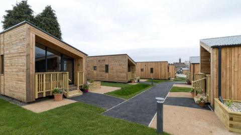 A housing village - two rows of small wood-panelled homes, facing each other, with grass and turf to walk on in-between them.