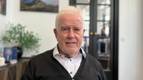 Roger Trower is a man with white hair, wearing a white shirt and a black cardigan. The background is an out-of-focus office.