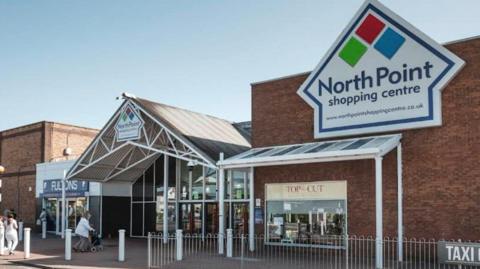 The image shows the main entrance of North Point Shopping Centre. Above the entrance is a large sign displaying the name 'North Point' alongside a logo made of three diamonds in red, green, and blue. The building has glass doors and windows. Two shops are visible: 'Fultons' on the left and 'Top Cut' on the right. A person is pushing a trolley near the entrance and there is a silver metal fence running across outside.