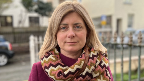 The picture shows a person standing outdoors wearing a deep burgundy coat and a thick patterned scarf with zig‑zag designs in shades of pink, brown, cream and green. Their hair is shoulder‑length and blonde. Behind them there is a metal fence, a pavement and parked cars, along with light‑coloured buildings in the background. 