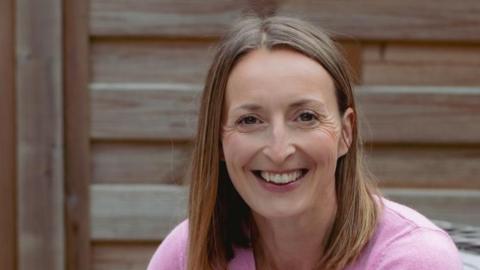 Close up of Caroline, in a pink jumper, smiling. There is a bare wooden fence behind her.