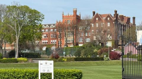 A large, grand brick school building, set behind a manicured lawn and tennis courts.