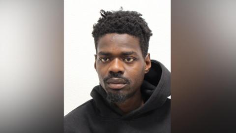 A police head and shoulders photo of a young man with a black beard and black hair, wearing a black hoodie. He is looking slightly off camera. 