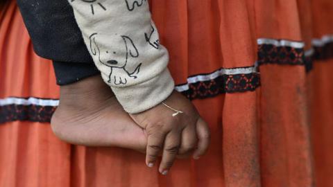 A close-up of a small child’s bare foot and hand is shown against a backdrop of their mother's skirt - a traditional indigenous outfit. It appears the child is being held by their mother. The child is wearing a light-coloured sleeve printed with a simple cartoon dog, and the hand hangs loosely beside the foot.