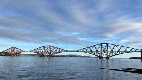 A wide view of the Forth Bridge across an expanse of sea under a cloudy, blue sky.