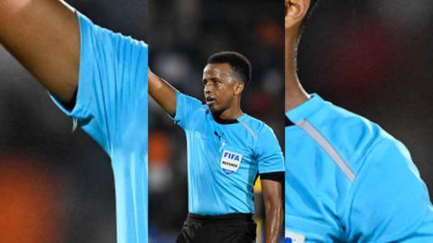 Omar Artan is wearing a light blue referee kit, and has his right arm in the air
