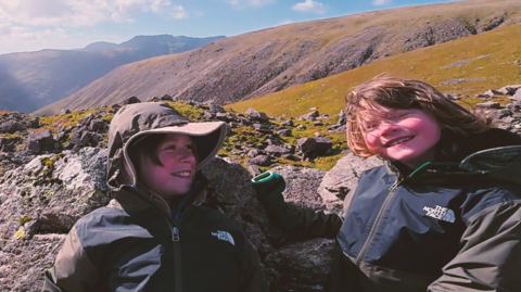 Two young boys smiling while sat on rocks on Scafell Pike in hiking gear 