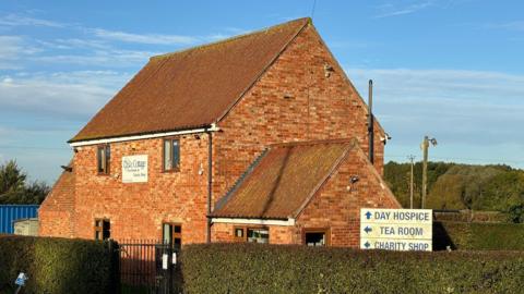 Two storey house with side building is the Dove Cottage tea room with hedges surrounding it and sign to hospice, tea room and charity shop