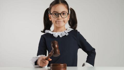 A young girl with brown pigtails wears judges clothing holding a gavel.