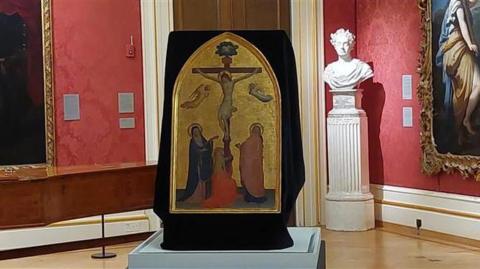 The Crucifixion with the Virgin, Saint John the Evangelist and the Magdalen by Fra Angelico in a room at the Ashmolean Museum in Oxford. A sculpture and other paintings can be seen behind it.