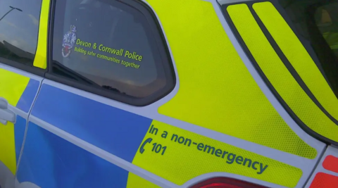 The side of a police vehicle. It has the words "Devon and Cornwall Police" on the window and is yellow and blue. 