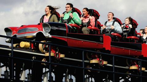 A rollercoaster with the design of a 1950s style American-style sedan speeds along a track. Several men, women and children are sat on the ride with stunned faces and hair blowing back in the wind.