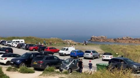 Cars in a sandy-gravel car park at Grandes Rocques, with the sea in the background