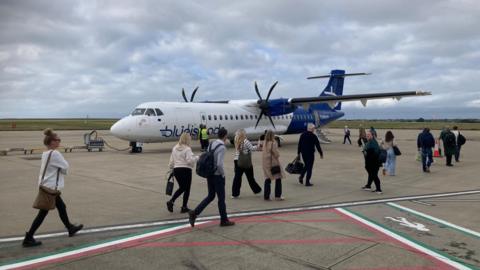 Passengers boarding a Blue Islands flight from Jersey to Birmingham 14 October 2024. The aircraft is mostly blue and white. Passengers are carrying their luggage while walking to the back end of the aircraft where they will board the plane. It is a grey day. 