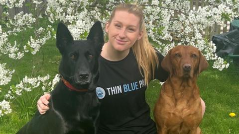 Kristen, a woman, wearing a black branded T-shirt, with her two dogs - a black one and another brown one, sitting either side of her outside on some grass, with white blossom on a tree behind her.
