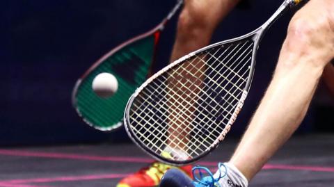 Generic image of close-up of squash racquet heads with white squash ball being hit, and two players' right legs visible