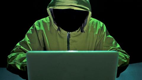 A hacker, wearing a green coat, against a black background stealing passwords and identity on a laptop.