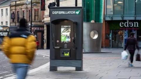 A high street showing a row of shops and a communication hub in the middle of the pavement. It looks like a dark grey phone box and says "defibrillator" on the top. The blurry figures of two people are walking past