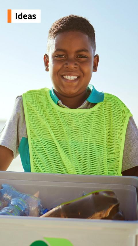 Young boy wearing a bright vest holding a box of plastic bottles