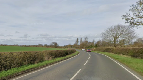 A country road with white lines down the middle and at the edges, with a small verge and low hedges on both sides. 