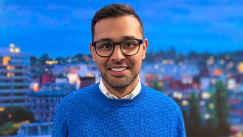Dan Miller looking and smiling at the camera with a blurred skyline of a city. He's wearing glasses and a blue jumper over a white shirt.