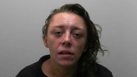 A custody photo of a woman sitting against a plain light‑coloured wall, wearing a black zip‑up jacket.