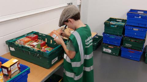 A child wearing a flat cap, a green t-shirt and an adult-sized work vest with Nuneaton Foodbank branding is labelling products from a green plastic crate. There are blue and green plastic crates stacked up in the room to the side of the child.