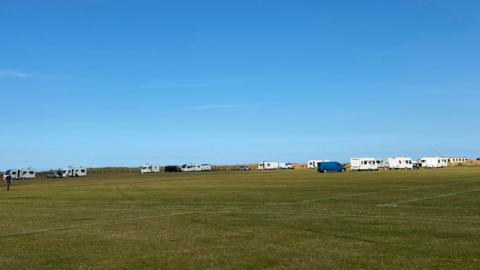 More than 10 white caravans have parked up along the edges of a football pitch. Two vans, one grey and one navy, are parked with the caravans. There are tyre marks in the grass heading towards where the camp has been set up.