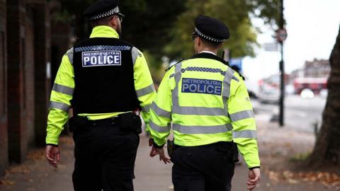 Two Metropolitan Police officers patrol a street in hi-vis jackets. Their backs are turned towards the camera. 