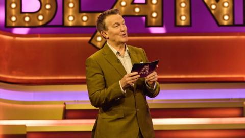 Bradley Walsh presents Blankety Blank. He is wearing a tan green suit.