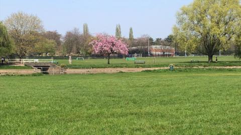 St Nicholas Park in Warwick. Photo shows the leisure centre in the background and a tree with pink cherry blossom as well as a white bridge and green football goal posts. A grass field is at the front of the picture. 