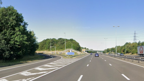 A three-lane motorway stretching away from the camera with cars driving on it. Across the central reservation is the other carriageways with cars and a lorry on it. Further down the road is a bridge running over it. In the foreground is a motorway junction lane splitting off and heading in a slight incline around a a left hand bend. There is a blue sign with white writing that reads "services"