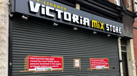 A shop front with a black shutter and a sign that reads 'Victoria Mix Store'. The shutter has a red notice stuck to it with the words 'This business has been closed by court order due to persons involved in the supply of illegal tobacco products". 