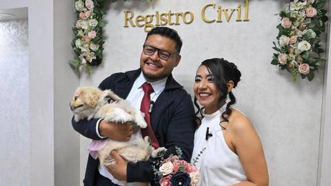 Andres and Diana pose with flowers, as Andres holding Luna who is dressed in a pink dress and bow.