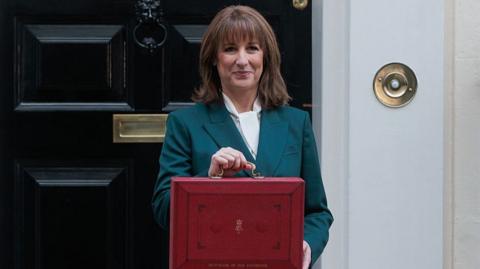 Reeves wears a green suit with a white shirt. She is holding a red briefcase in front of her. 