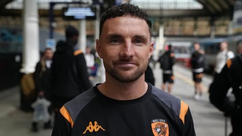 Lewie Coyle standing in front of other players on the platform at Hull Station.
He has short dark hair and a beard and wears a black and amber Hull City T-shirt
