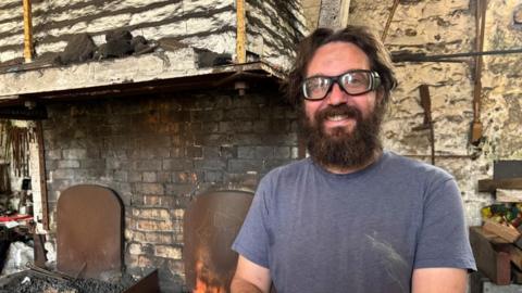 A bearded man stands in front of the camera, he's wearing a dirty blue t-shirt. He is wearing safety goggles and is smiling. Behind him to the right is a blacksmith fire and a soot-covered wall.