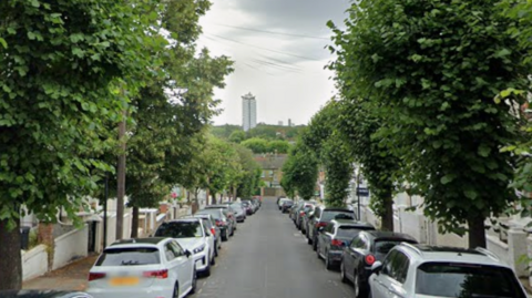 A Google Maps image of a road lined with houses and trees and a large tower block in the distance.