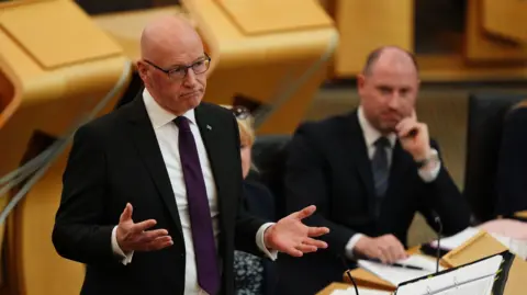 John Swinney, wearing a dark suit, white shirt and purple tie, holds his hands in front of him as he answers questions at Holyrood