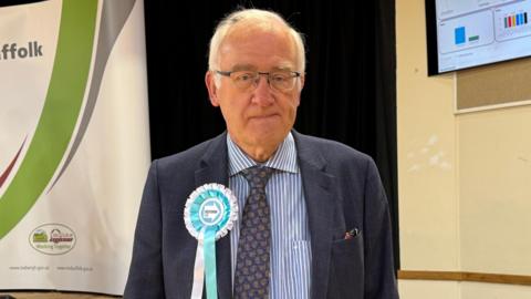 A man with grey hair and glasses smiles at the camera. He wears a navy suit with a white and blue striped shirt. There is a light blue and white rosette pinned to his chest. 