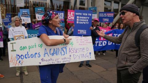 A doctor holding a sign opposing assisted dying legislation in front of supporters of the law as she argues with a man.