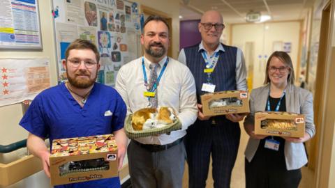 The image shows four people standing in a hospital hallway, smiling and holding boxed fake pets labelled "Precious Petzzz." The man on the far left is wearing blue hospital scrubs. He has short ginger hair and a ginger beard and moustache. The man on the right of him is wearing grey suit trousers, a white shirt and a grey patterned tie. The man to the right of him is wearing dark blue striped suit trousers, a dark blue striped waistcoat and a light blue shirt. The woman on the far right is wearing a black outfit with a white blazer. A dementia awareness board is visible in the background on the left. 