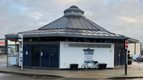 A former bus station. It is a low, possibly octagonal building with a white wall, with a bicycle resting against it, and black doors. Its black roof rises into a pinnacle. Beneath the roof and on the white wall it says "Welcome to Mildenhall High Town".